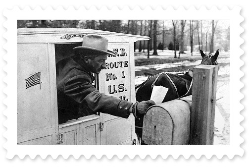 A black-and-white photograph depicting a postal worker leaning out of a horse-drawn carriage to place two letters in a rural-area mailbox.