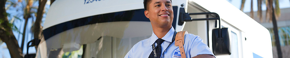 USPS mail carrier scanning a Priority Mail box while standing in front of a Next Generation Delivery Vehicle (NGDV).