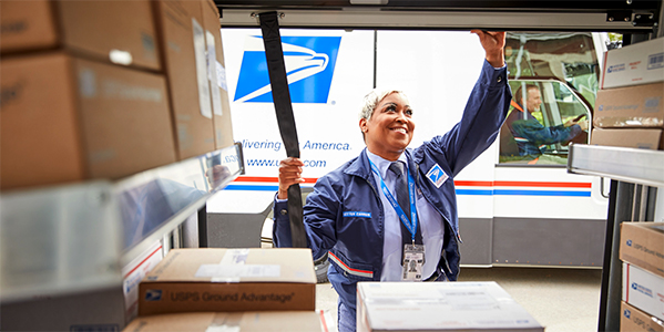 USPS mail carrier scanning a Priority Mail box while standing in front of a Next Generation Delivery Vehicle (NGDV).