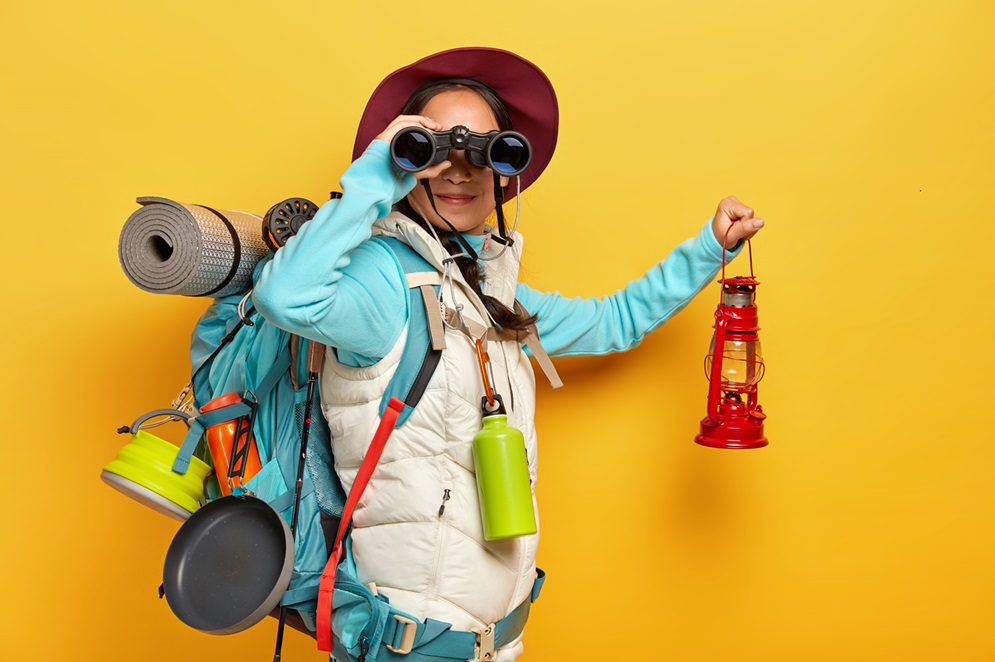 Horizontal shot of curious female backpacker explores tourist destination, uses binoculars, dressed in active wear, holds kerosene lamp carries travelling items with rucksack isolated over yellow wall